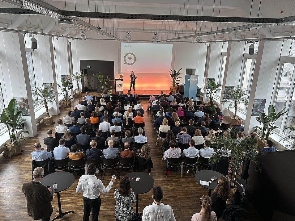 Fotografie in den großen Saal mit vielen Menschen, deren Aufmerksamkeit auf die Referentin auf der Bühne gerichtet ist