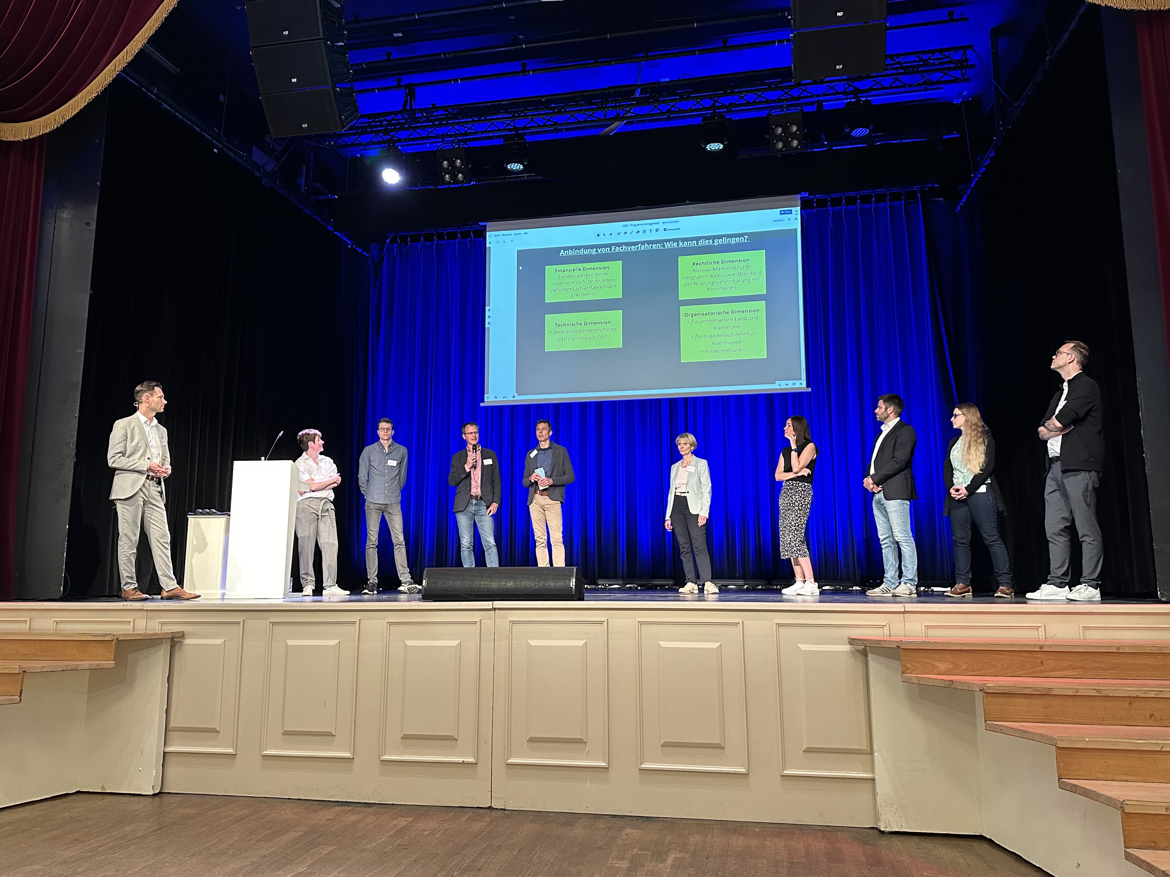 Referent:innen stehen auf einer Bühne, im Hintergrund eine Leinwand mit Vortragsfolien