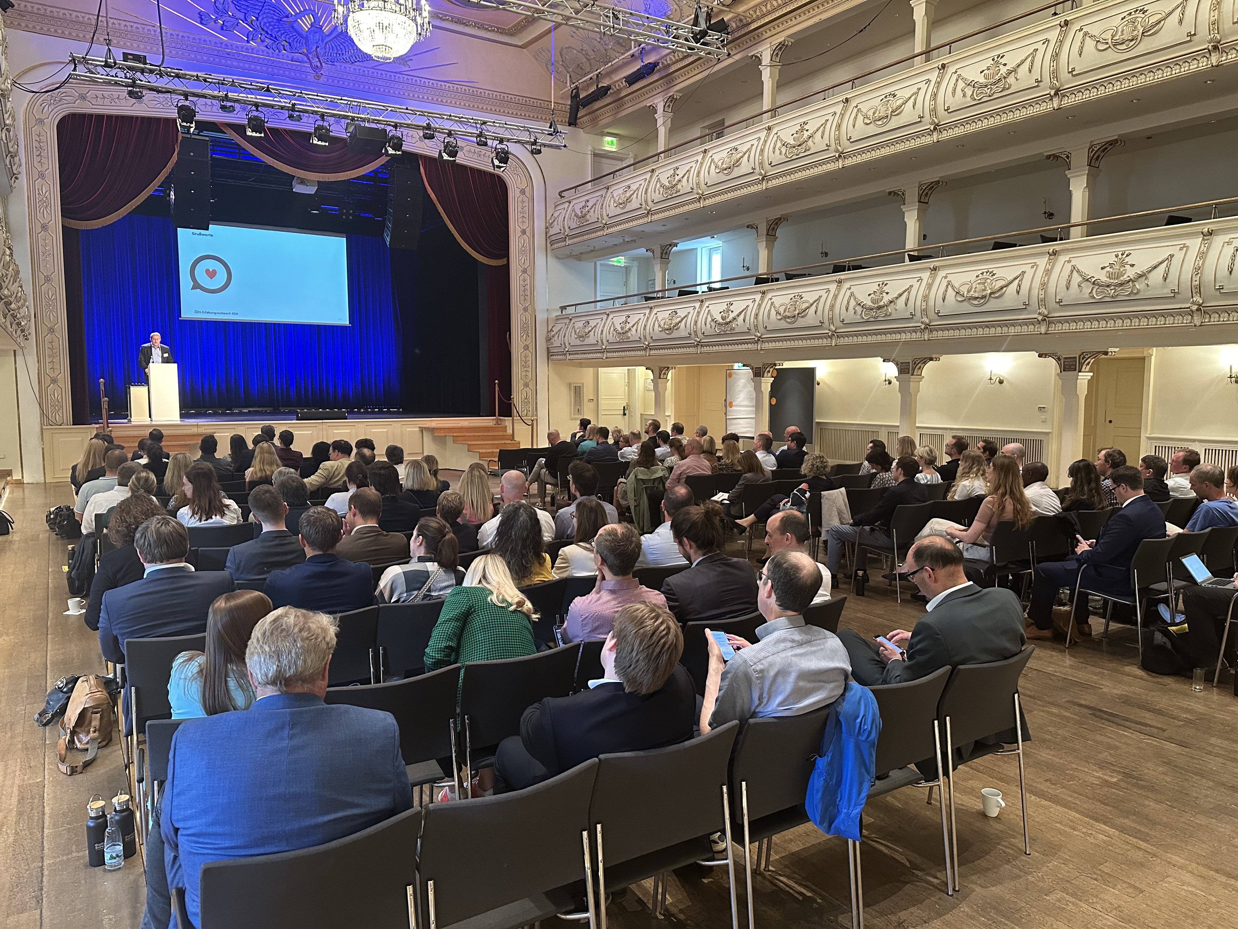 Saal voller Menschen in Stuhlreihen, die nach vorne auf eine Bühne blicken, auf der ein Moderator steht