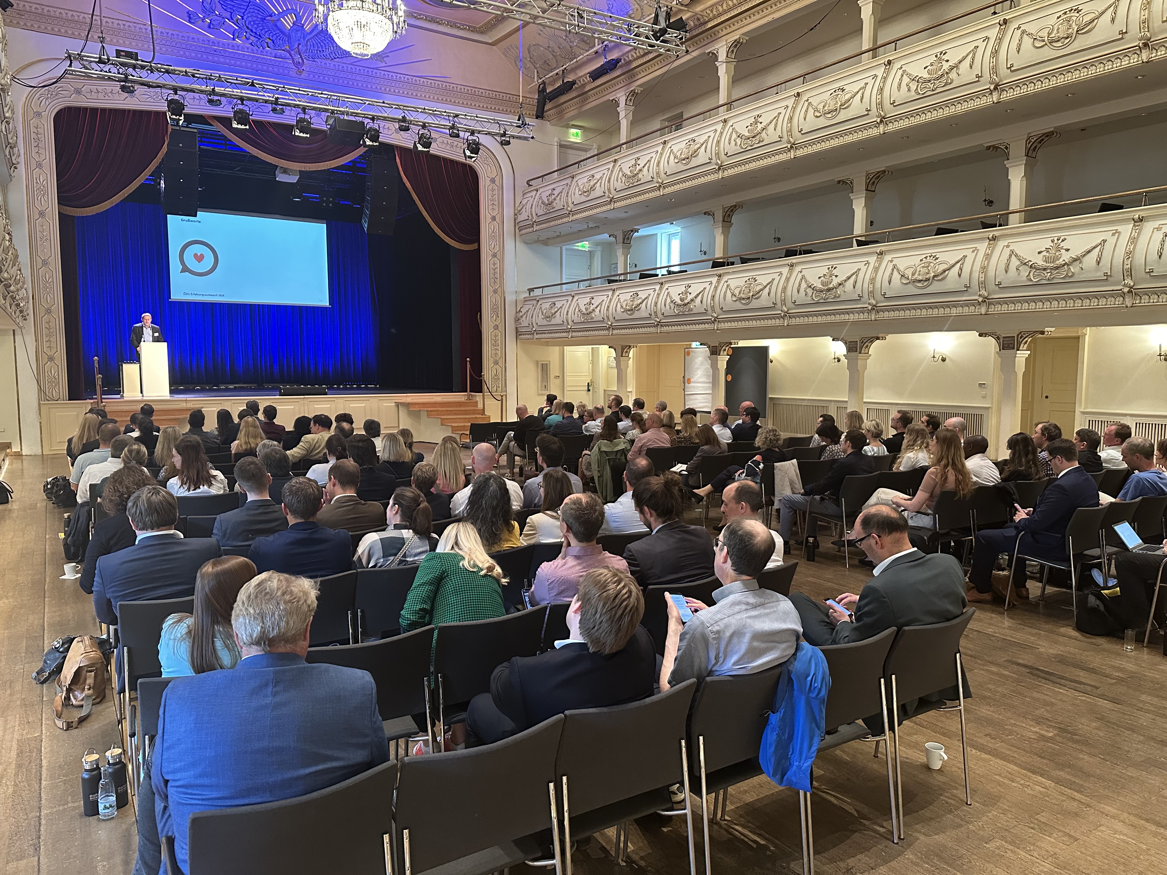 Saal voller Menschen in Stuhlreihen, die nach vorne auf eine Bühne blicken, auf der ein Moderator steht